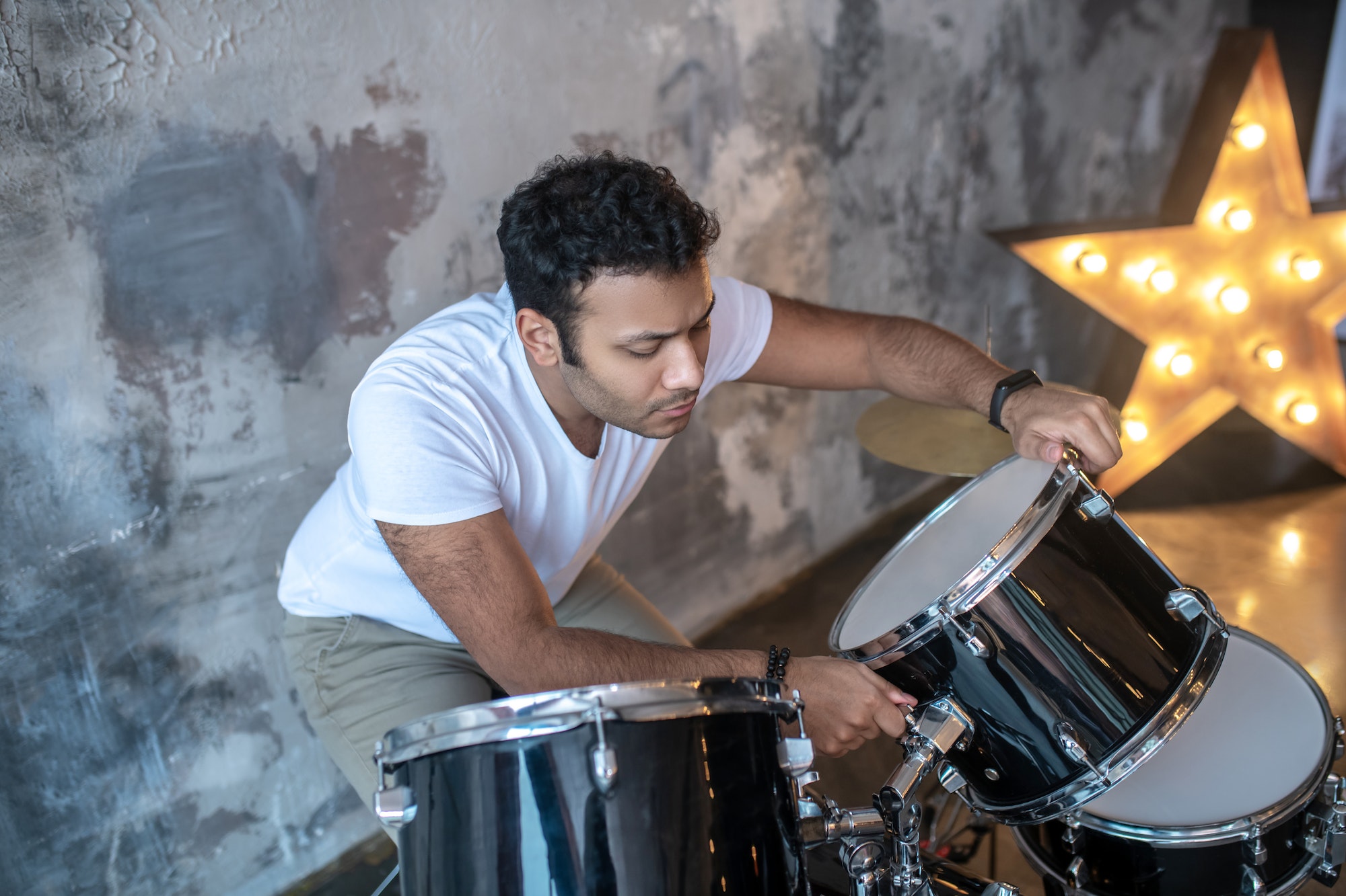 Dark-haired young man in a white tshirt standing near the drums